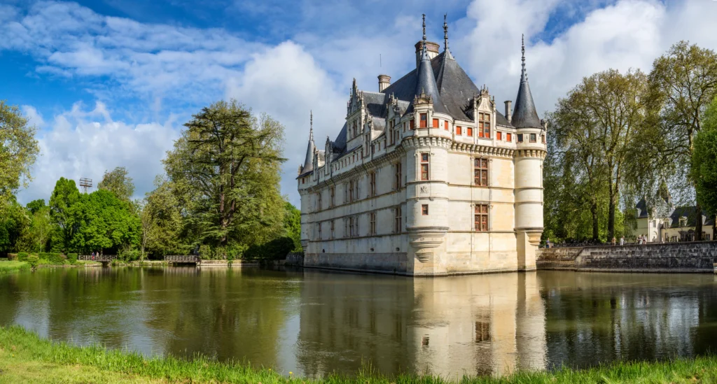 Vue du château d’Azay-le-Rideau se reflétant dans les eaux calmes de l’Indre, entouré d’arbres et de verdure.